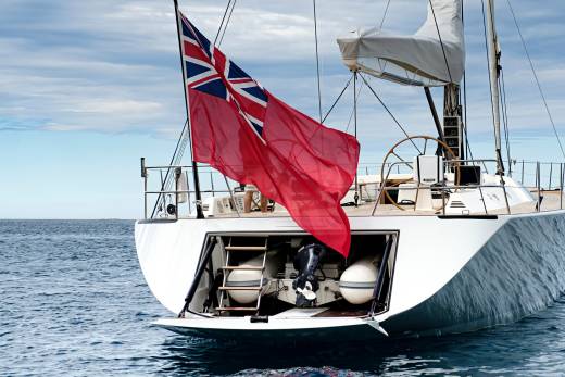A British Red Ensign flag flutters proudly from the stern of a sleek white yacht sailing on calm blue waters.