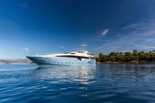A sleek, light blue yacht with a dragon motif glides through calm, blue waters near a tree-lined coast.