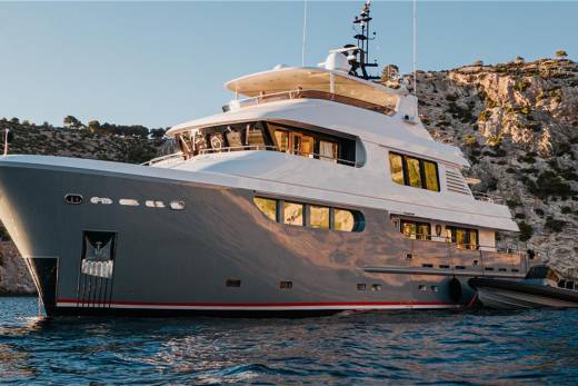 A luxurious yacht glides through calm blue waters near a rocky, tree-covered coastline.