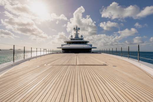 The sun glints off the vast wooden deck of a luxurious yacht as it sails through the sparkling blue ocean under a cloudy sky.