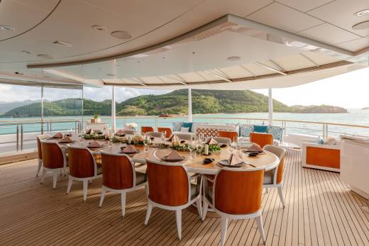 An elegant outdoor dining table is set for a feast on a yacht, overlooking a serene turquoise bay and lush green hills.