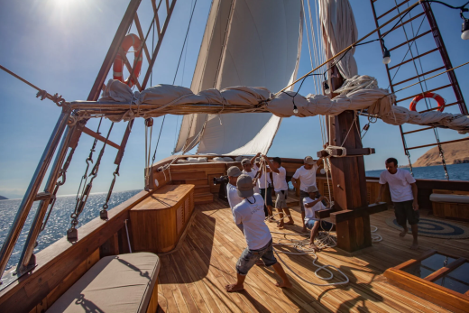 A crew works together to hoist the sails on a traditional wooden schooner under a bright blue sky.