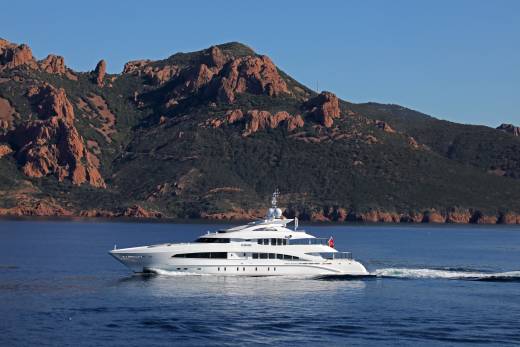 A sleek white yacht glides through the azure waters with a dramatic red-rock mountain range providing a stunning backdrop.