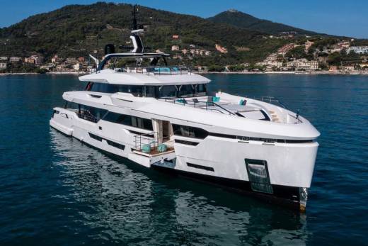 A sleek white yacht glides through the calm blue waters with a picturesque coastal town nestled on the green hillsides in the background.