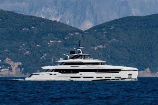 A sleek white yacht glides through the deep blue sea with a lush, mountainous coastline in the background.