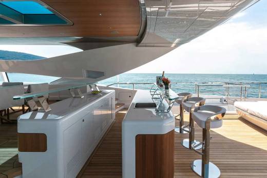 Luxurious yacht deck features a sleek bar with stools, dining area, and sun loungers overlooking the serene ocean.