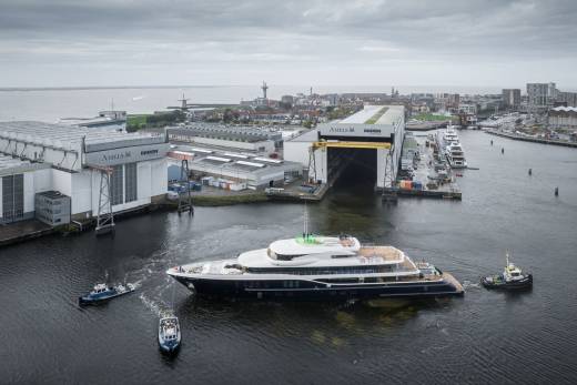 A massive yacht is guided out of a shipyard by two tugboats under a cloudy sky.
