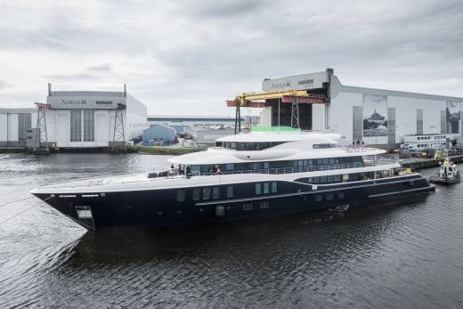 A sleek, dark-hulled superyacht glides from its shipyard under an overcast sky.