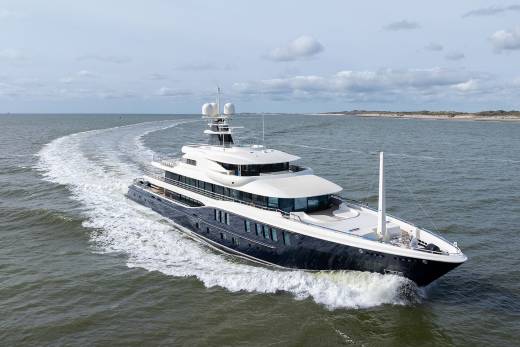 A sleek, dark-hulled superyacht cuts through the choppy water, leaving a white wake against a backdrop of dunes and sky.