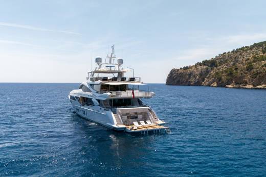 A luxurious white yacht cruises through the deep blue Mediterranean Sea near a rocky, tree-covered island.