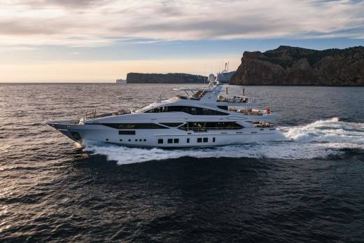 A sleek white yacht cuts through the dark blue sea, leaving a trail of white foam against a backdrop of rugged cliffs and a soft sunset sky.