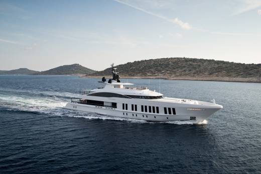 A sleek white superyacht cruises through the deep blue waters of a Mediterranean island chain under a clear sky.