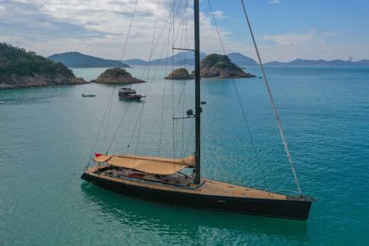 A sleek black sailboat with a tan sail glides through the turquoise waters of a scenic bay dotted with islands.