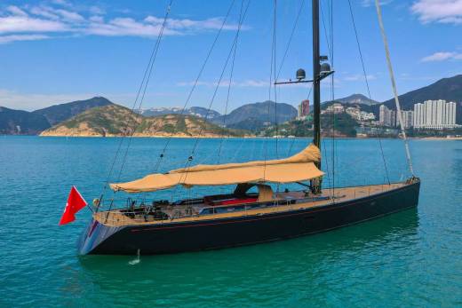 A sleek black yacht with tan sails glides through turquoise waters against a backdrop of lush green hills and a distant city skyline.