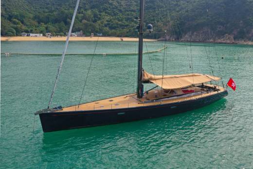 A sleek, dark sailboat with a wooden deck glides through turquoise waters near a sandy beach.