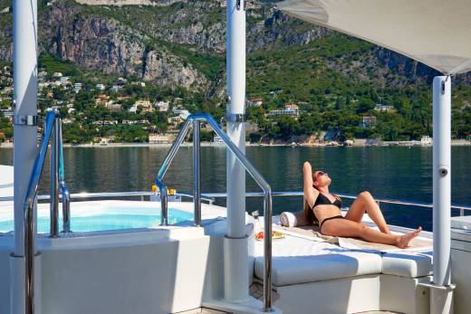 A woman sunbathes on a yacht's deck with a hot tub and a scenic coastal town in the background.