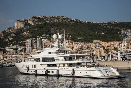 A luxurious white yacht, named "New Secret," is docked in a harbor with a densely populated hillside city in the background.