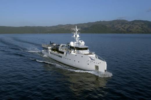 A sleek white superyacht cruises through calm blue waters with lush green mountains in the background.