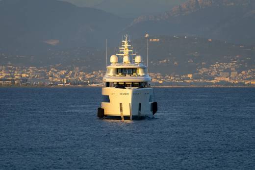 A luxurious white yacht glides across calm blue waters with a picturesque coastal city nestled at the base of distant mountains.