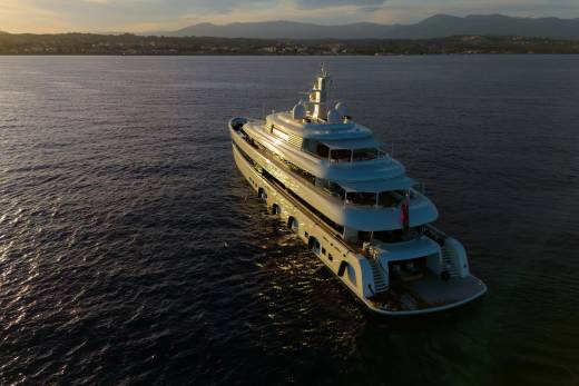 A luxurious white yacht glides across calm waters as the sun sets behind a distant coastal town.