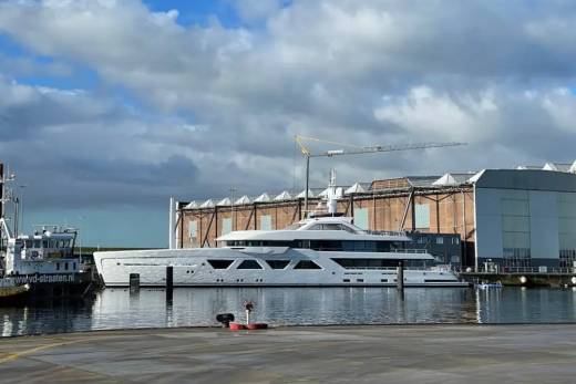 A sleek, modern superyacht is docked at a shipyard with a large industrial building and crane in the background.
