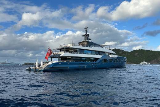 A large, luxurious yacht with a sleek, modern design sits in the calm, blue waters, surrounded by lush, green hills and a picturesque cloudy sky.