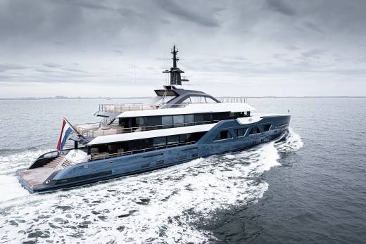 A large, sleek yacht with multiple decks and a tall mast is speeding across the choppy, gray waters of what appears to be an open sea or lake, with a cloudy, overcast sky in the background.