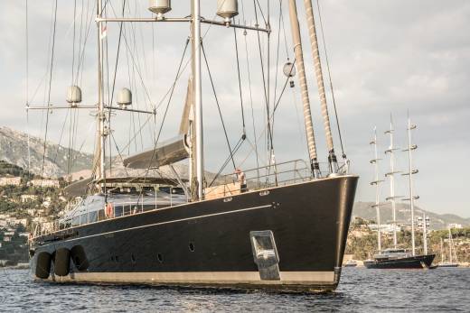 A sleek black yacht anchors offshore with a majestic three-masted schooner sailing in the background.