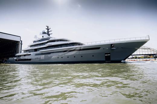A massive, sleek grey yacht is docked in a shipyard, its modern design gleaming under a bright sky.