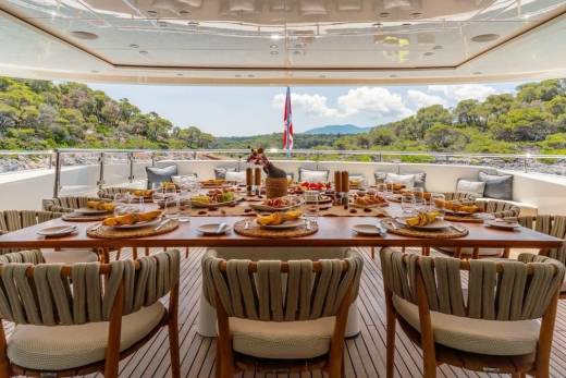 A beautifully set dining table awaits guests on a yacht deck, overlooking a lush, green coastline.
