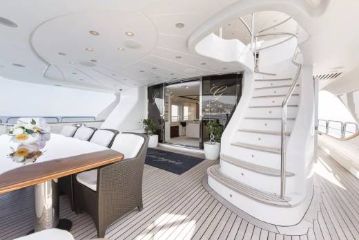 An elegant outdoor dining area on a yacht deck features a large table, comfortable seating, and a sweeping staircase leading to upper levels.