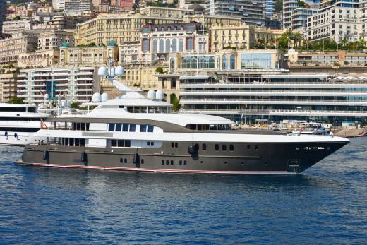 A sleek, dark-hulled superyacht glides through blue waters with a backdrop of Monaco's iconic cityscape.