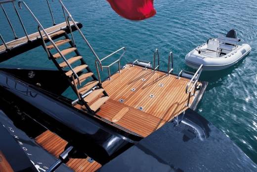 A luxurious yacht's teak deck and wooden stairs lead to the turquoise water where a tender awaits.