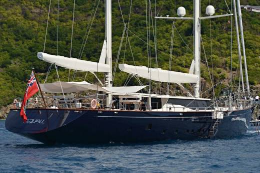 A majestic dark blue sailing yacht glides through calm blue waters with lush green hills in the background.