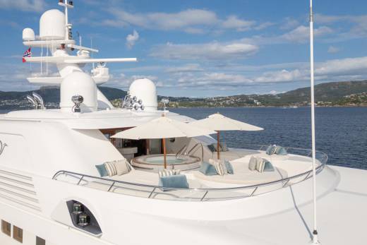 A luxurious yacht with a hot tub and sun loungers is anchored in a calm bay with a picturesque coastline.