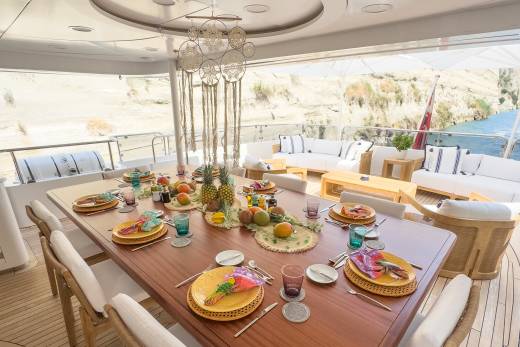 A beautifully set dining table on a yacht deck awaits a meal with a scenic backdrop of cliffs and water.