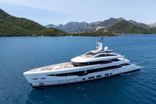A luxurious white superyacht glides through the calm, blue waters of a scenic bay, with lush green hills in the background.