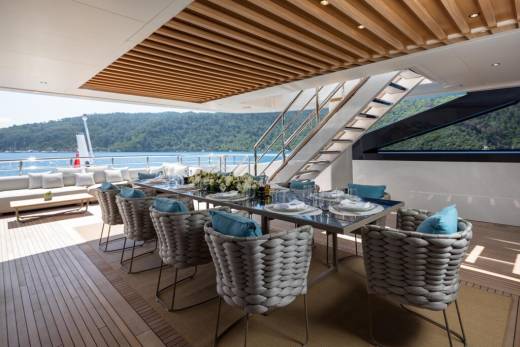 An elegant outdoor dining area on a yacht features a long table set for a meal with a backdrop of lush green hills and sparkling blue water.