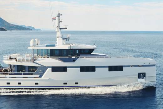 A sleek white yacht cuts through the calm blue sea, with a mountainous coastline visible in the distance.