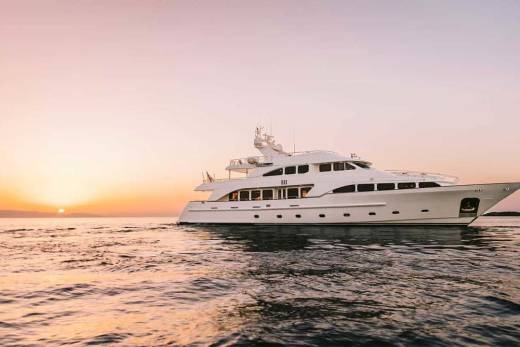 A luxurious white yacht glides across calm waters at sunset, silhouetted against a soft pink and orange sky.
