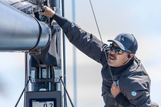 A sailor adjusts rigging on a boat, his face obscured by sunglasses and a cap.