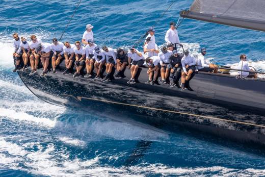 A team of sailors leans over the dark hull of a racing yacht as it cuts through the choppy blue ocean.