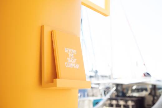 A bright yellow book titled "Beyond the Yacht Company" is displayed on a matching shelf against a blurred backdrop of yachts.