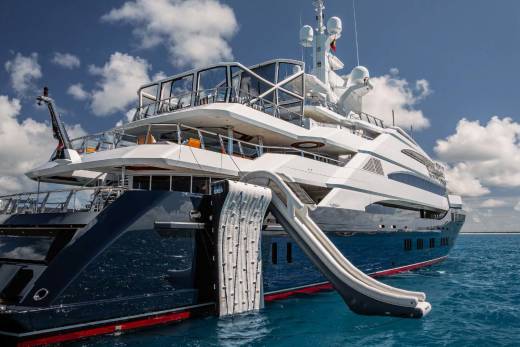 A large, luxurious white yacht with multiple decks and a slide is anchored in the calm, blue waters, with a cloudy sky in the background.