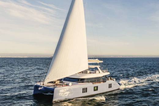 A large white sailboat with a tall mast glides across the calm blue waters, with a scenic horizon visible in the background.