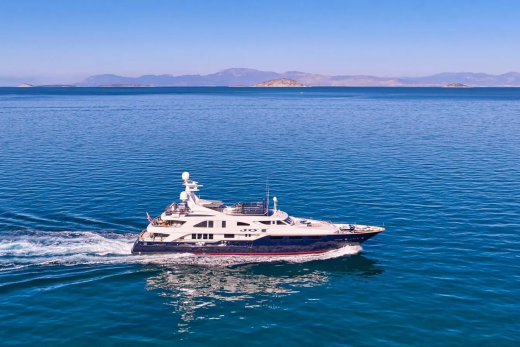 A sleek yacht cruises across the azure waters, leaving a white wake against the backdrop of distant, hazy mountains.