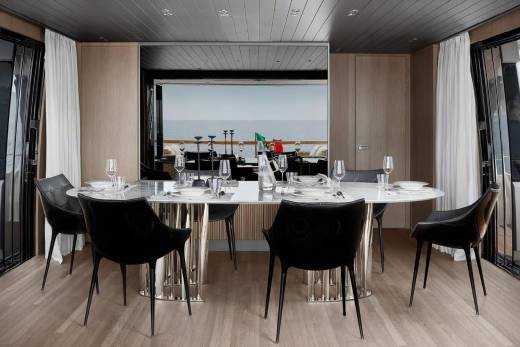 A modern and sleek dining area with a large table and black chairs, surrounded by large windows offering a scenic view of the ocean in the background.