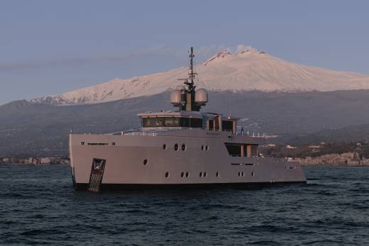 A large, gray yacht is floating on the calm waters of a bay, with a snow-capped mountain peak visible in the background.