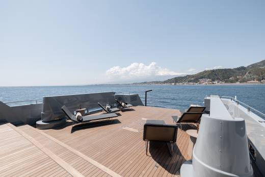 A wooden deck overlooking a scenic coastal landscape, with mountains and buildings in the background and a calm body of water in the foreground.