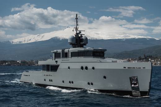 A large, sleek white yacht navigates the calm waters, with snow-capped mountains visible in the background against a cloudy sky.